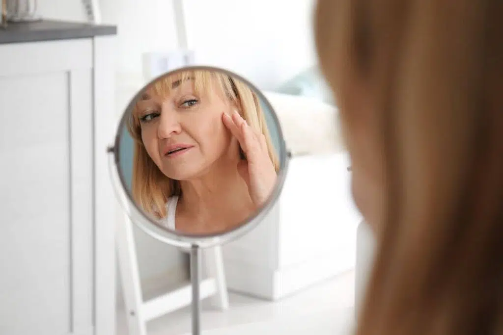 Woman examining sagging facial skin in mirror after weight loss, showing early signs like jowls and hollow cheeks.