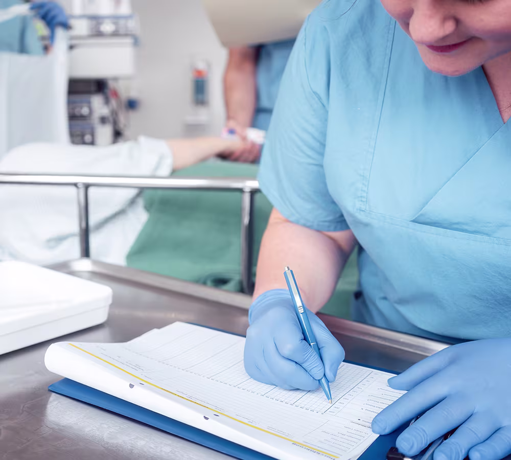 Nurse in blue scrubs reviewing a pre-op checklist beside a patient bed before revision rhinoplasty procedure