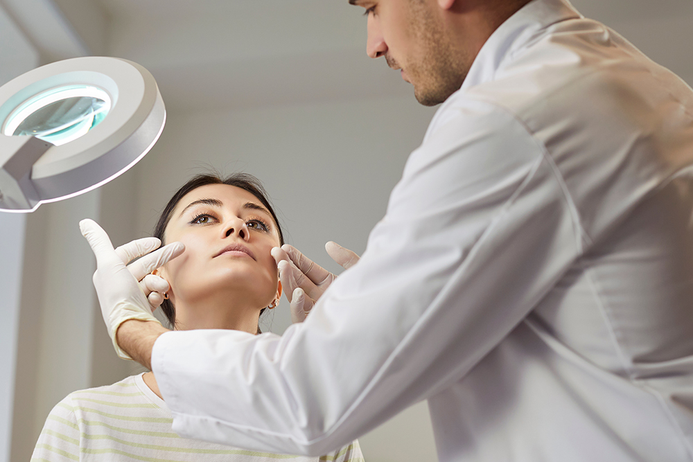 Doctor assessing female patient’s face under magnifying light before facial fat transfer to improve cheek and jawline volume