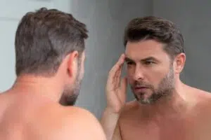 A man with a short beard looking into a bathroom mirror and using his hand to examine the fine lines and skin laxity around his eye and temple.