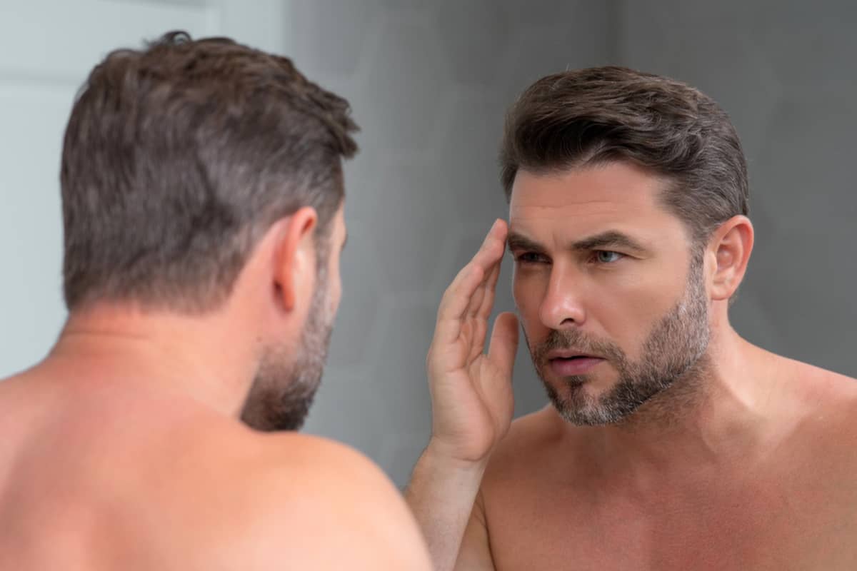 A man with a short beard looking into a bathroom mirror and using his hand to examine the fine lines and skin laxity around his eye and temple.