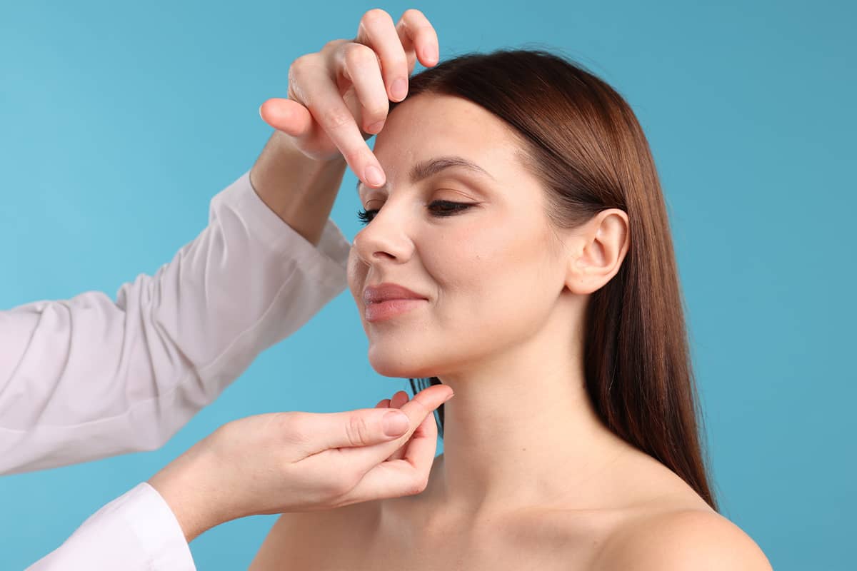 A surgeon in a white lab coat gently touching a woman's forehead and chin to evaluate her facial structure and skin elasticity.