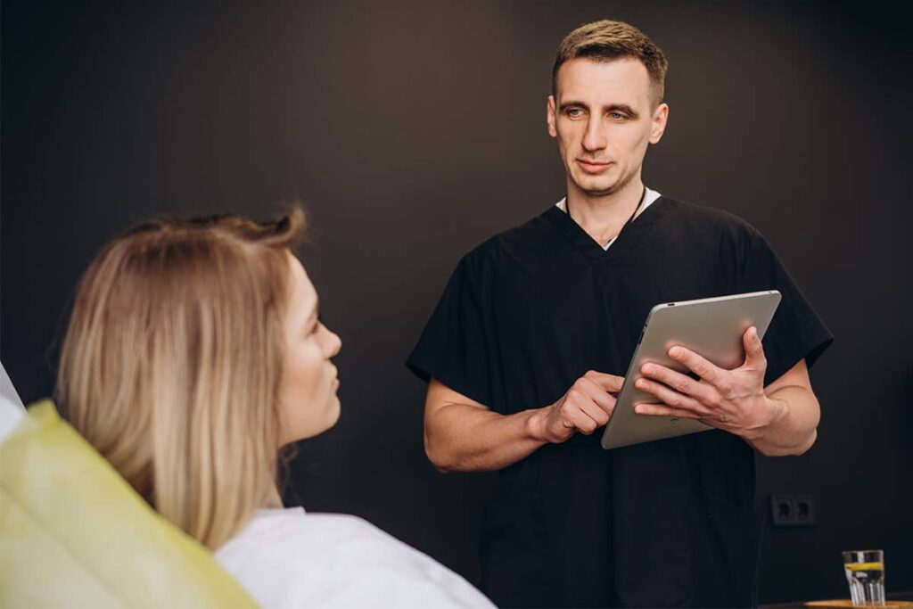 A male surgeon in black scrubs holding a tablet while discussing treatment options with a patient during a medical consultation.
