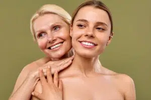 A younger woman and a middle-aged woman with glowing skin smiling together against an olive green background.