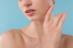 A close-up view of a woman’s lower face, lips, and neck against a blue background as she gently touches her smooth jawline.