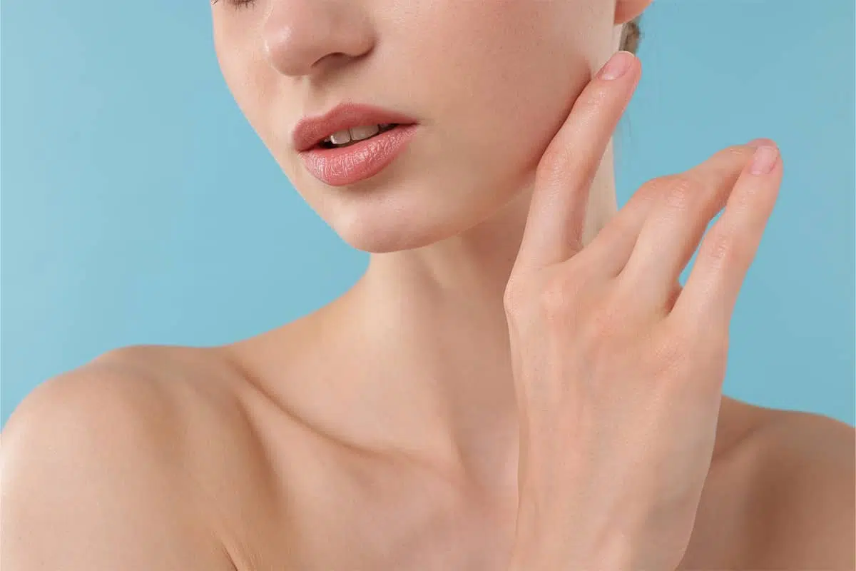 A close-up view of a woman’s lower face, lips, and neck against a blue background as she gently touches her smooth jawline.